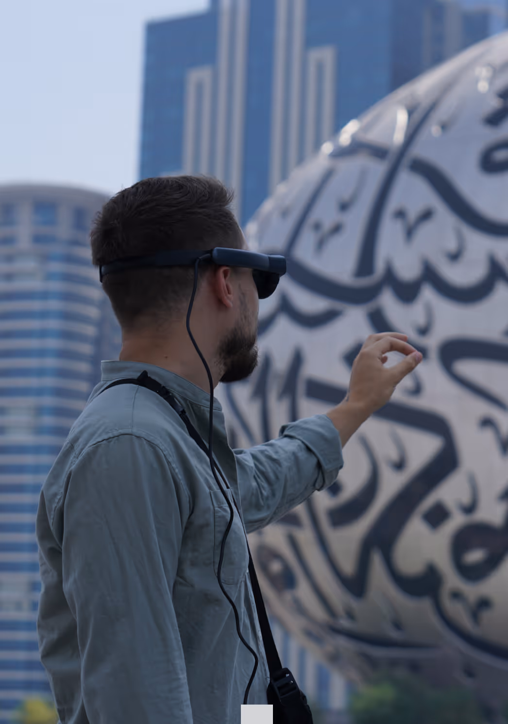 Man wearing augmented reality glasses reaching toward a large spherical sculpture with Arabic calligraphy in an urban setting.
