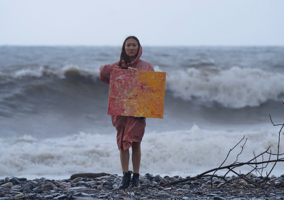 Woman in a pink hooded coat holding an abstract painting with red and yellow colors standing on a rocky beach with ocean waves in the background.