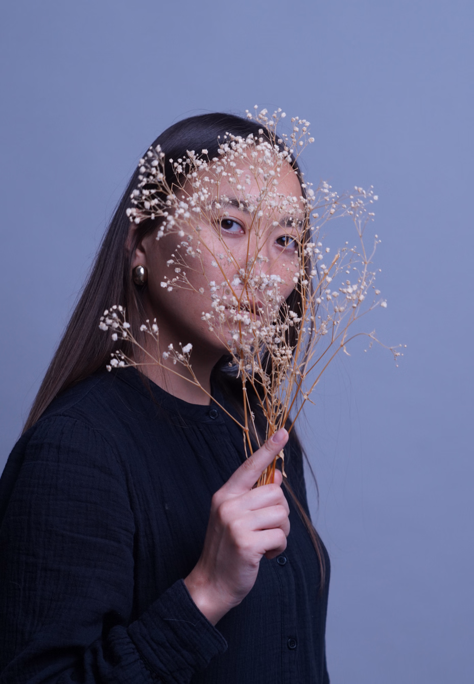 Woman in a black shirt holding delicate white baby's breath flowers in front of her face against a plain blue background.