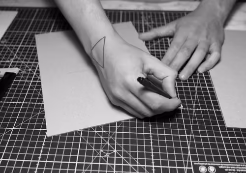 Person sketching on a piece of cardboard on a grid cutting mat, with a pen in hand and a triangle tattoo on the wrist.