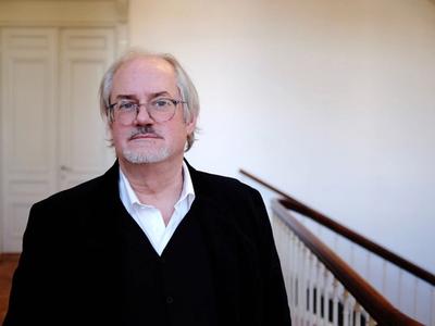 Portrait of an older man with glasses and white hair wearing a black suit standing indoors near a wooden handrail staircase.