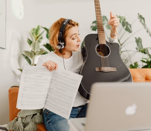 Woman with headphones holding a black acoustic guitar and music sheet while sitting on a couch with a laptop in front of her.