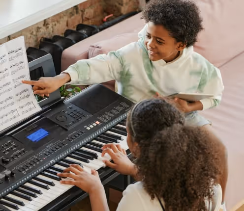 Child pointing at sheet music while another child plays an electric keyboard piano.