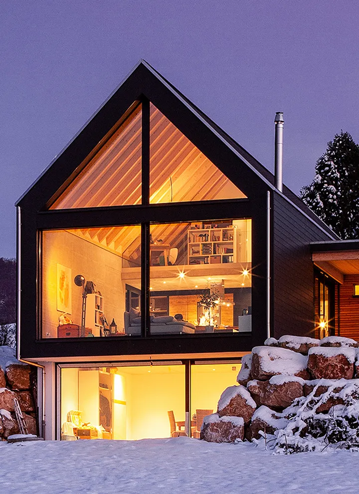 Maison moderne avec grandes fenêtres illuminées montrant un intérieur chaleureux en bois, entourée de neige et de rochers.