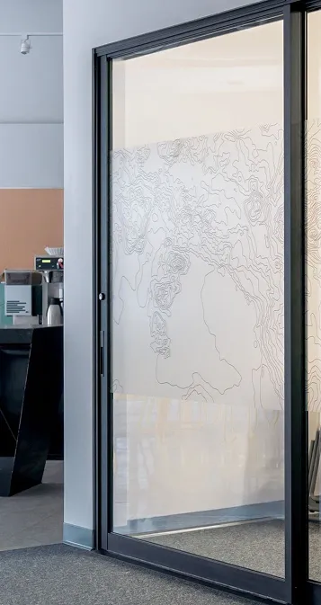 Porte en verre avec un film décoratif à motifs de contour topographique dans un bureau moderne avec une machine à café en arrière-plan.