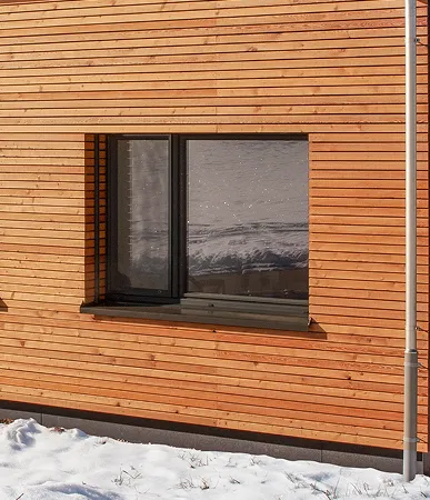 Fenêtre avec cadre noir sur mur en bois clair, avec neige au sol et reflet de paysage enneigé dans la vitre.