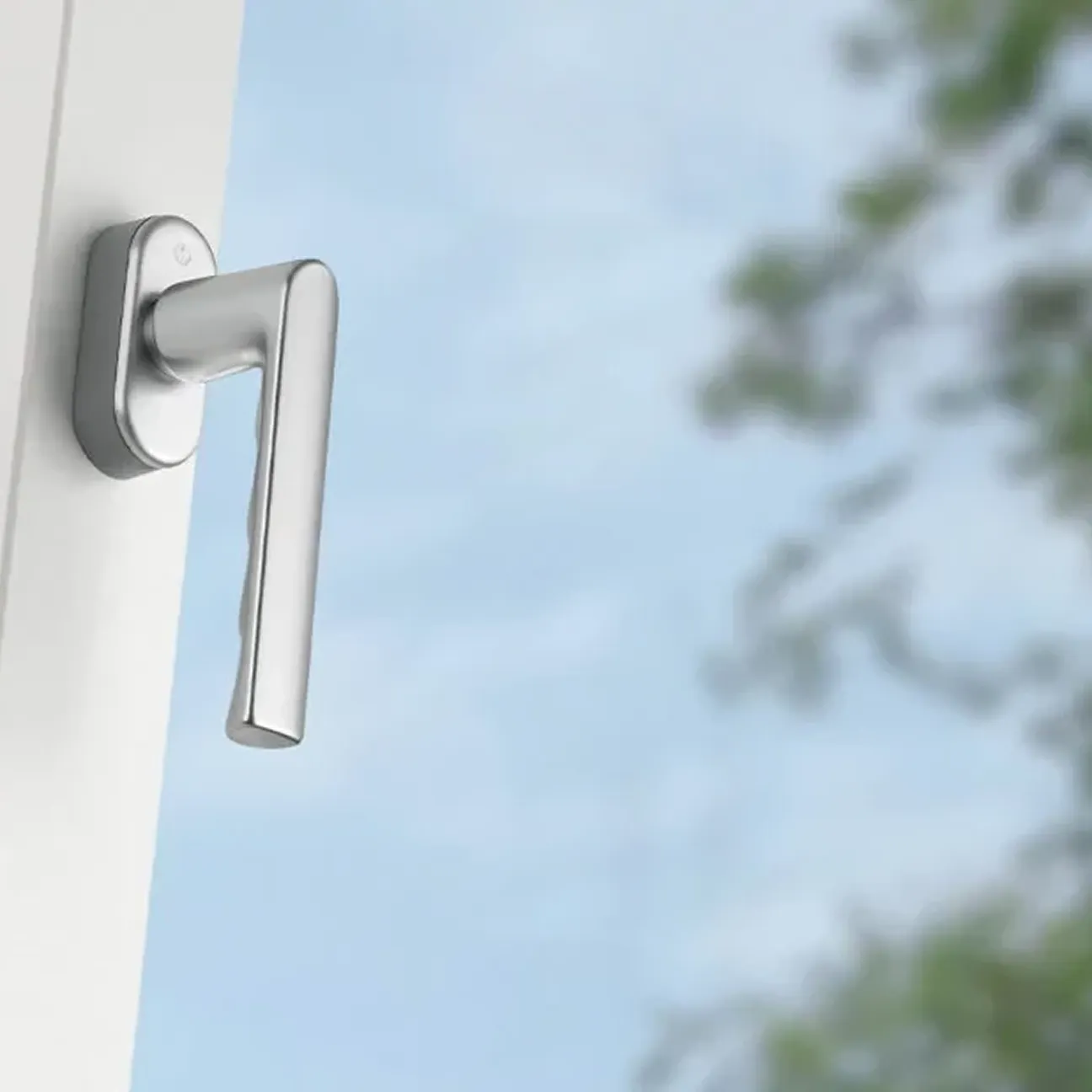 Ein silberfarbener Fenstergriff an einem weißen Fensterrahmen vor dem Hintergrund eines blauen Himmels und von Laub.