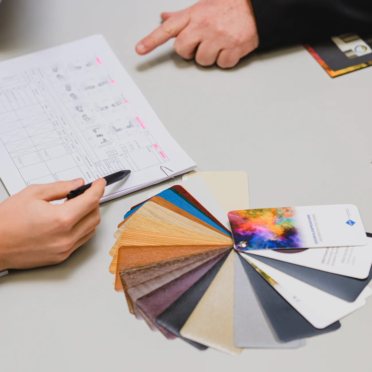 Zwei Personen unterhalten sich vor einer Materialfarbkarte und technischen Plänen, die auf einem Tisch liegen.
