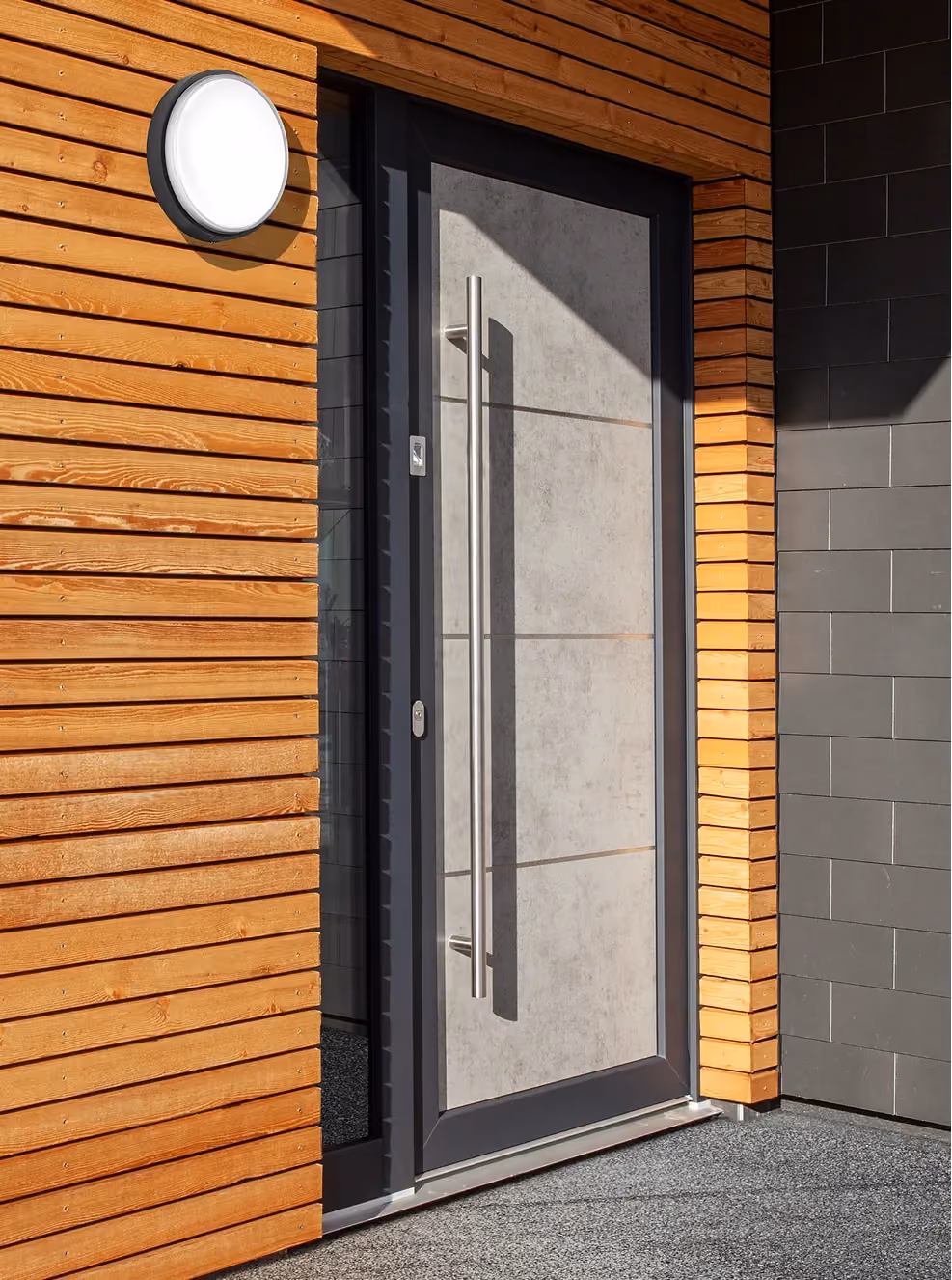 Entrée moderne avec une grande porte grise Domofen, un mur lambrissé et un luminaire extérieur rond. Design lumineux et contemporain.