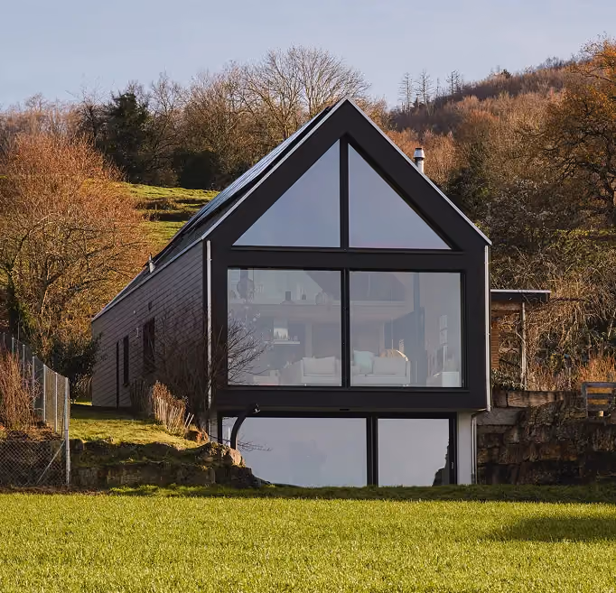 Maison moderne suisse avec une grande façade vitrée, se détachant sur un fond d'arbres et de collines, entourée de pelouse et d'une clôture.