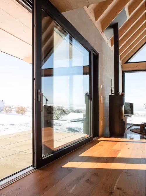 Porte coulissante en verre avec cadre noir donnant sur une terrasse enneigée, intérieur en bois clair avec lumière naturelle.