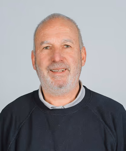 Portrait d'un homme âgé souriant avec une barbe grise et portant un pull noir.