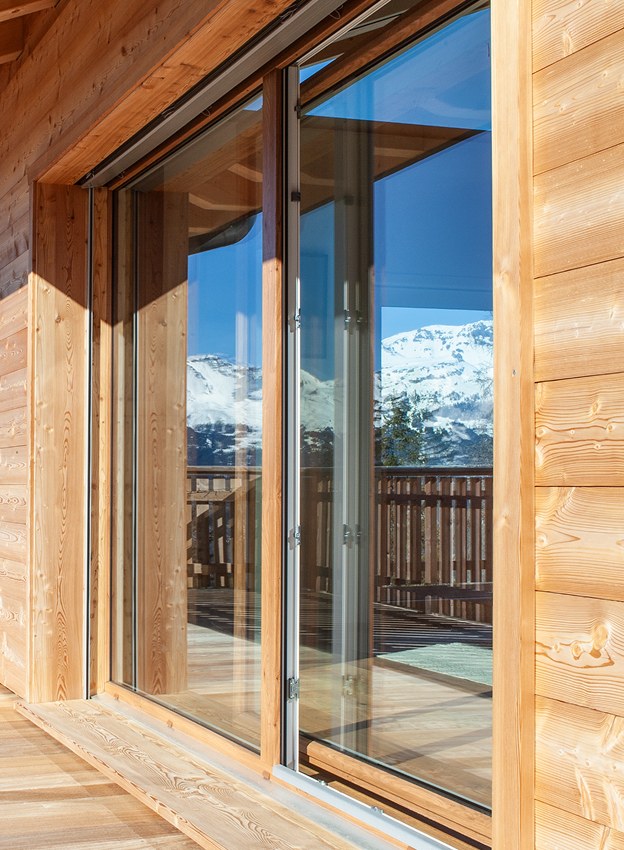 Fenêtre coulissante en bois avec réflexion des montagnes enneigées et d'une terrasse en bois.