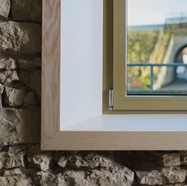 Nahaufnahme eines hellen Holzfensterrahmens mit weißer Innenseite vor einer Wand aus Naturstein, in der sich draußen eine Brücke und Bäume spiegeln.