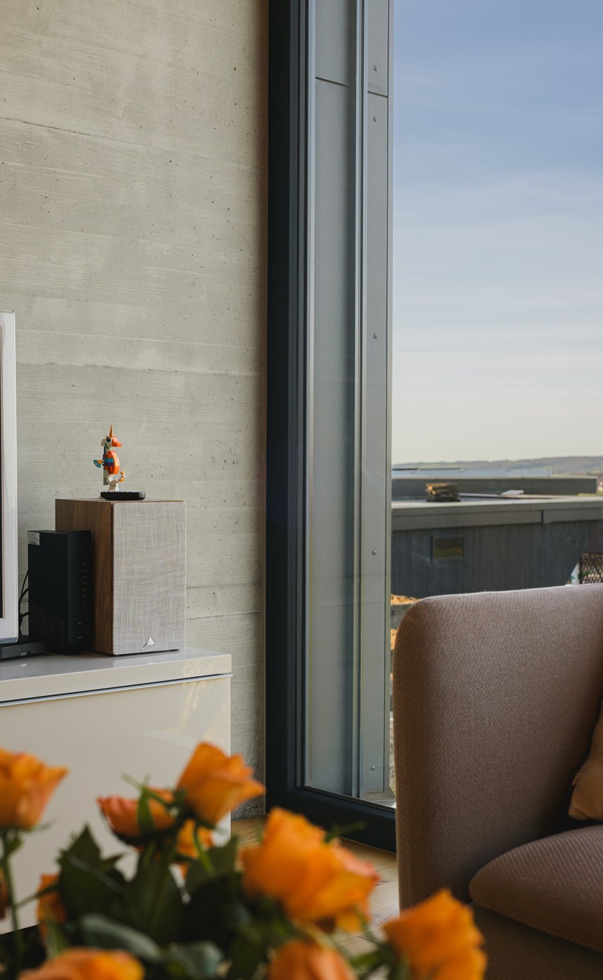 Intérieur lumineux avec un fauteuil beige près d'une grande fenêtre donnant sur un balcon, avec une enceinte et une figurine sur un meuble contre un mur en béton.