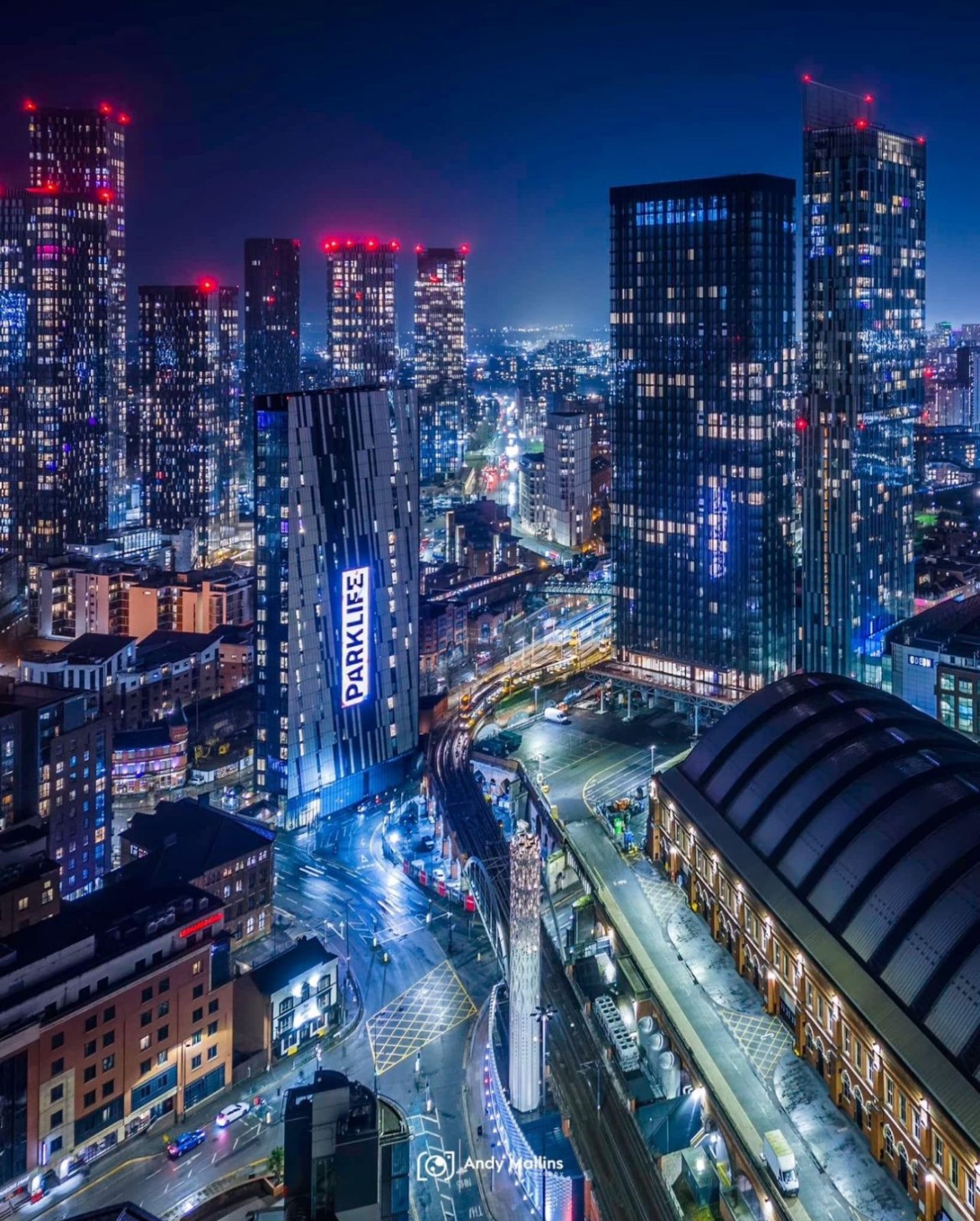 Nighttime aerial view of a cityscape with high-rise buildings lit up, including a tilted building displaying a bright 'PARK LIFE' sign and illuminated streets below.