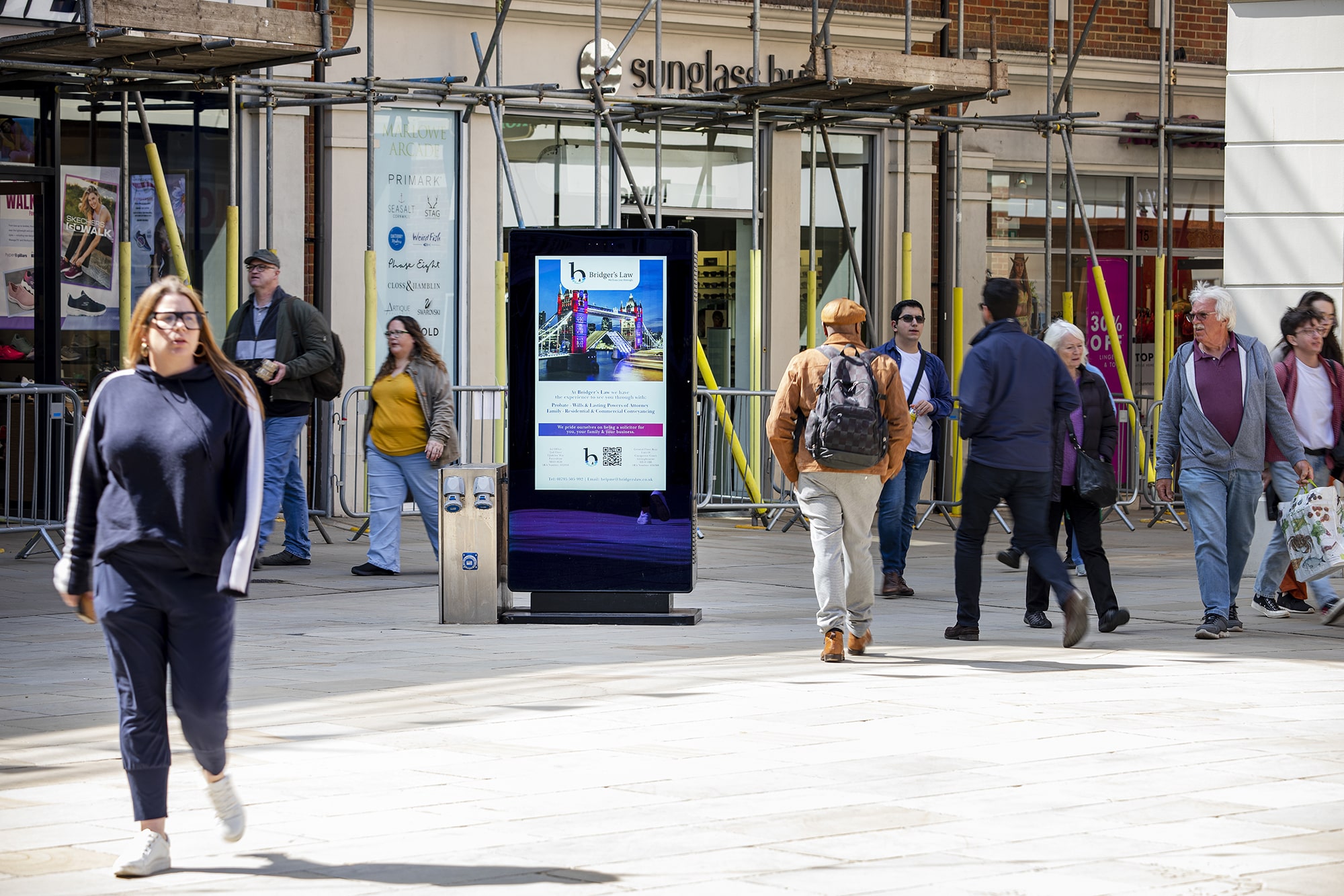 People walking on a city street near a digital advertisement display in front of a sunglasses store.