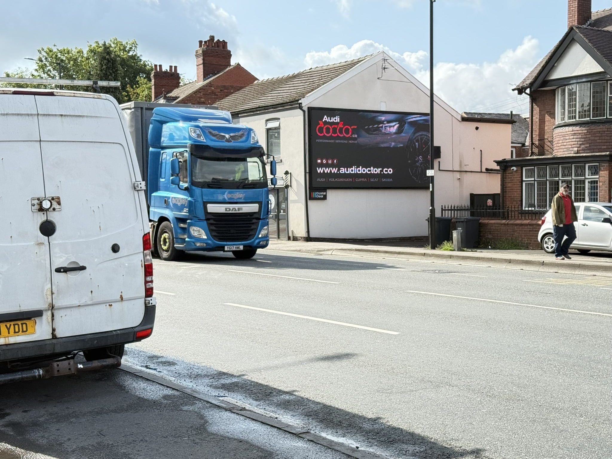 Street view with a blue DAF truck, a white van, a man walking on the sidewalk near a house, and a large AudiDoctor advertisement on a building wall.