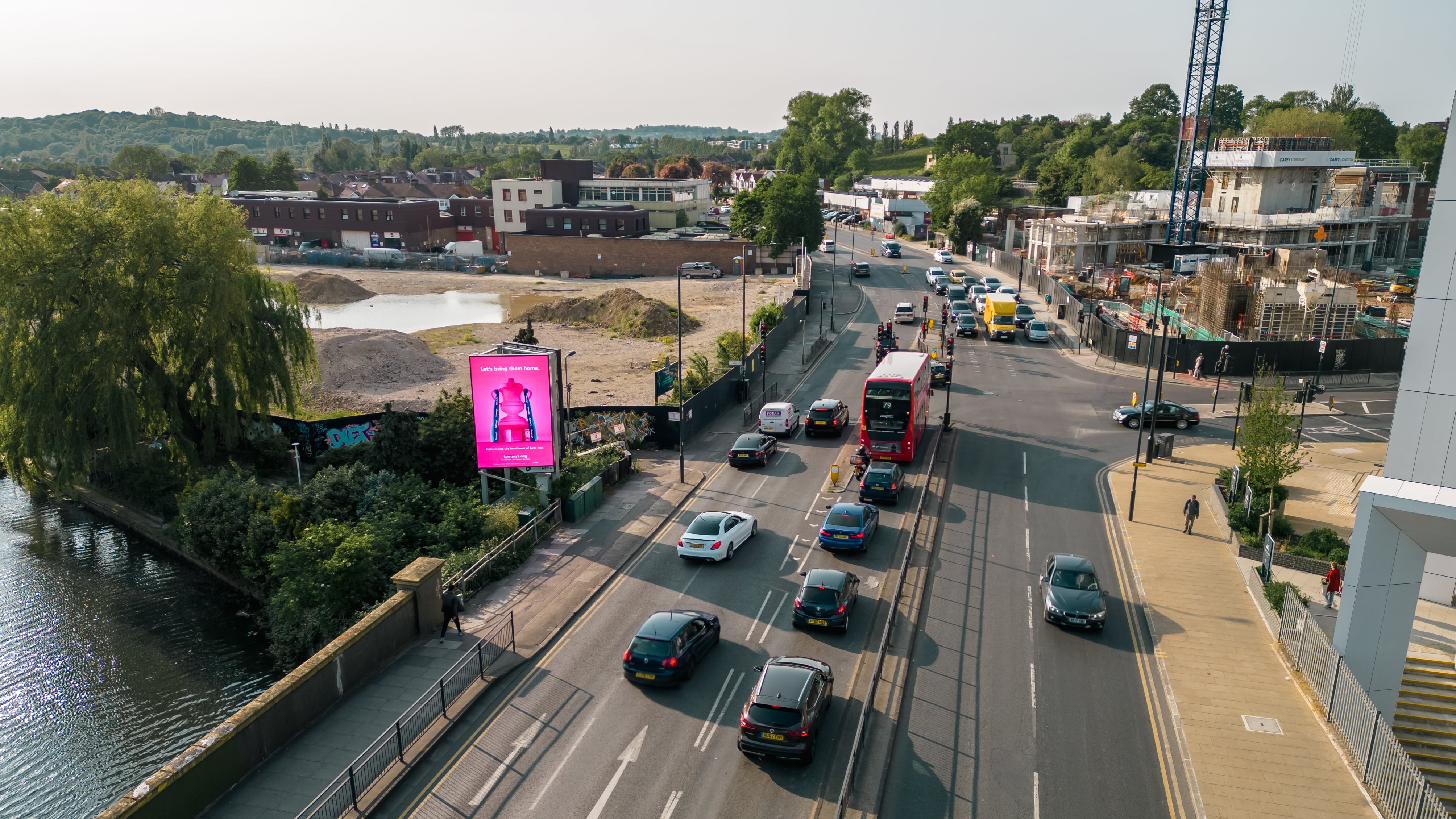 Aerial view of a busy urban road with multiple cars, a red double-decker bus, a river on the left, construction site on the right, and greenery in the background.