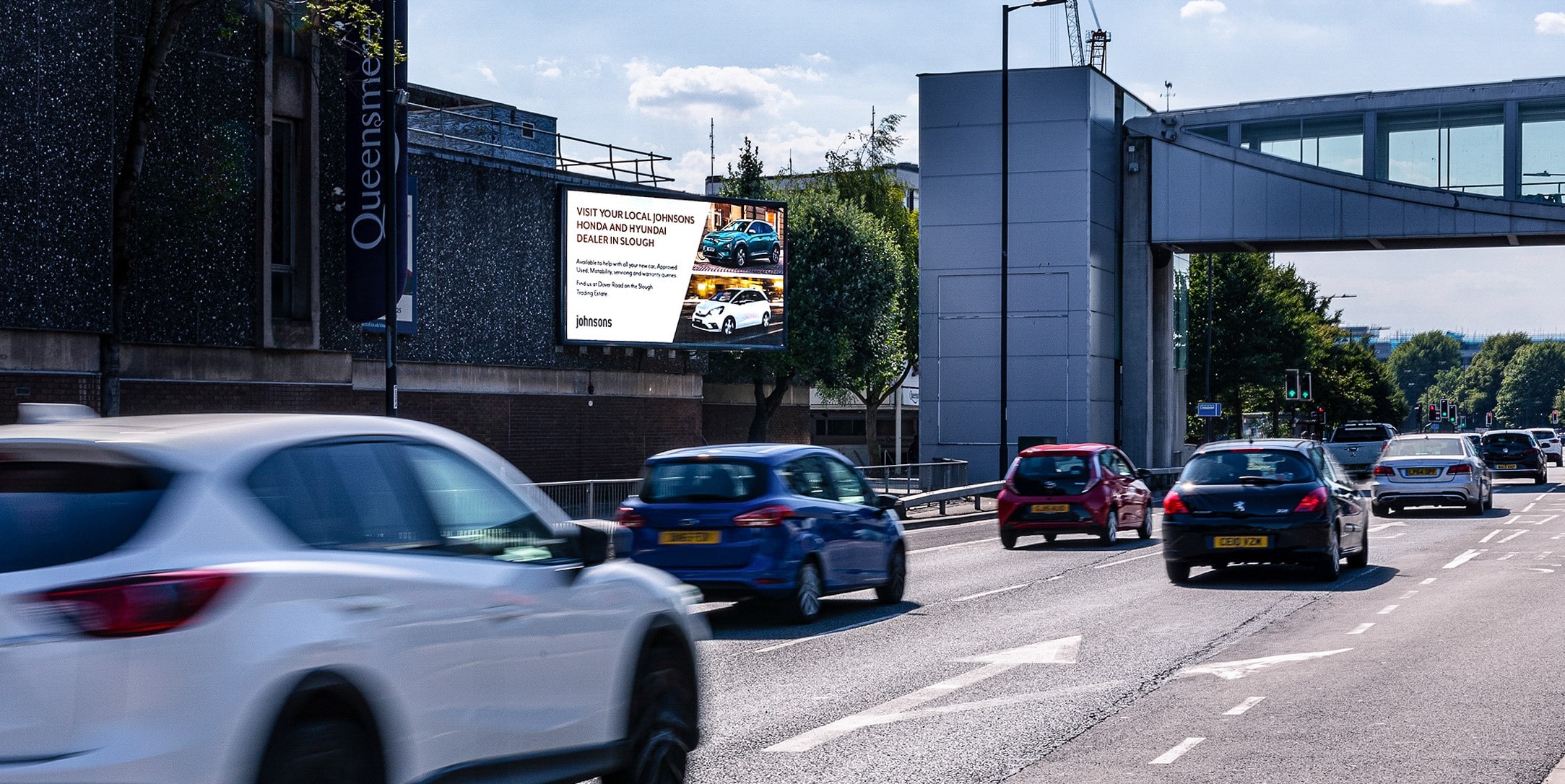 Busy urban street with multiple cars driving under a pedestrian bridge and a large digital billboard advertising Johnsons Honda and Hyundai dealership.
