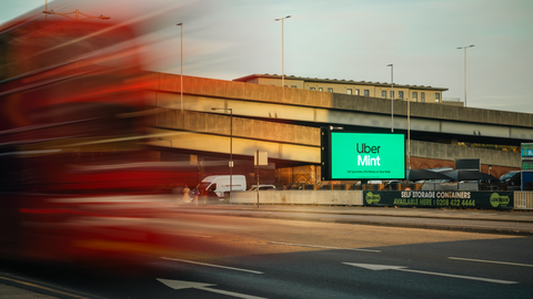 Blurred red double-decker bus passing on road in front of overpass with digital Uber Mint advertisement.