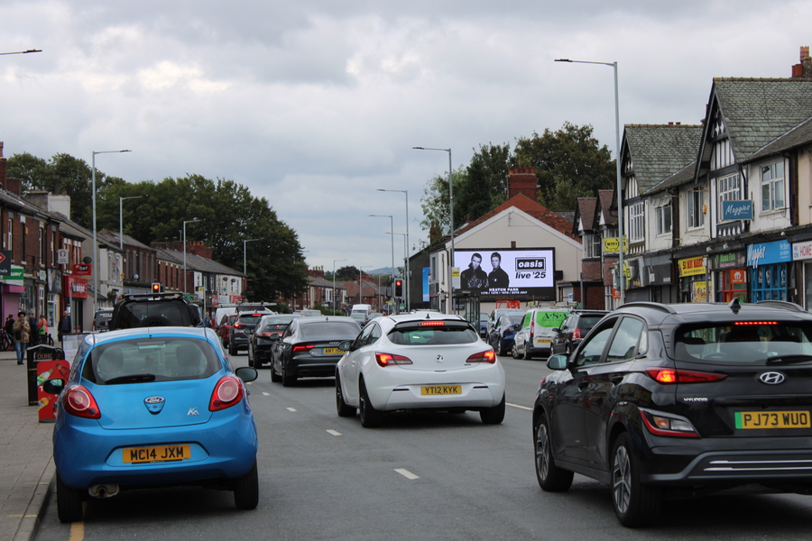 Busy street with several cars stopped at a traffic light, lined with brick and Tudor-style buildings under a cloudy sky.