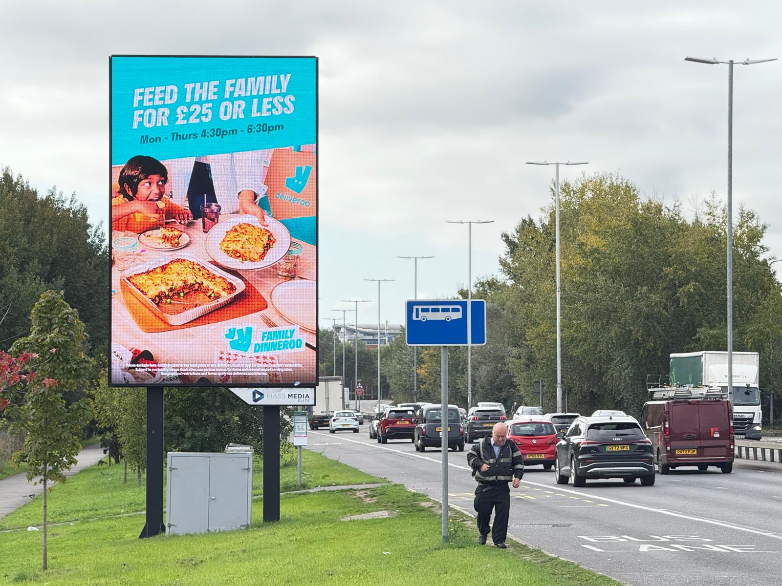 Digital billboard on roadside advertising Deliveroo family dinner offer for £25 or less, with traffic and pedestrian near bus lane sign.