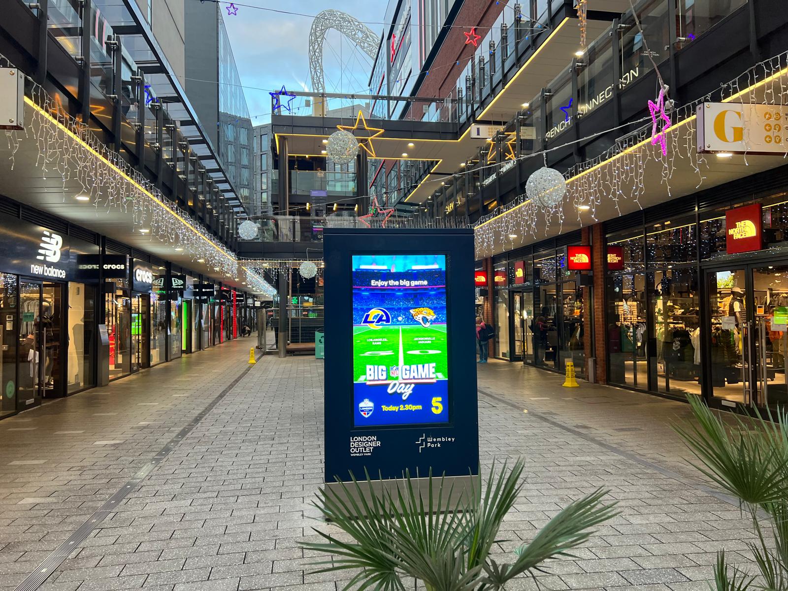 Indoor shopping mall corridor decorated with hanging star and orb lights, featuring a digital display advertising an NFL game between Los Angeles Rams and Jacksonville Jaguars.