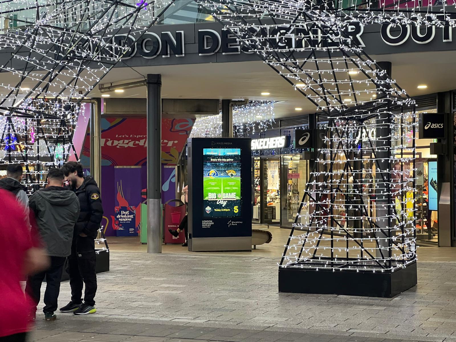 Outdoor shopping area at night with light decorations shaped like Eiffel Tower supports, people standing, and illuminated store signs including Skechers and Asics.