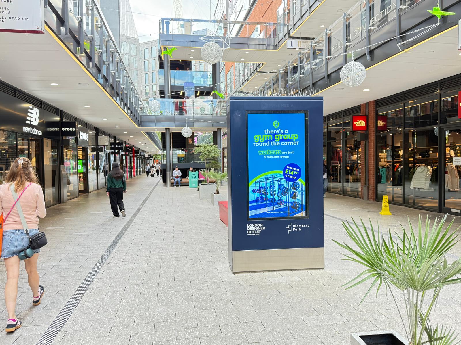Outdoor shopping area with people walking past stores like New Balance and The North Face, featuring a digital advertisement for a gym group at London Designer Outlet, Wembley Park.