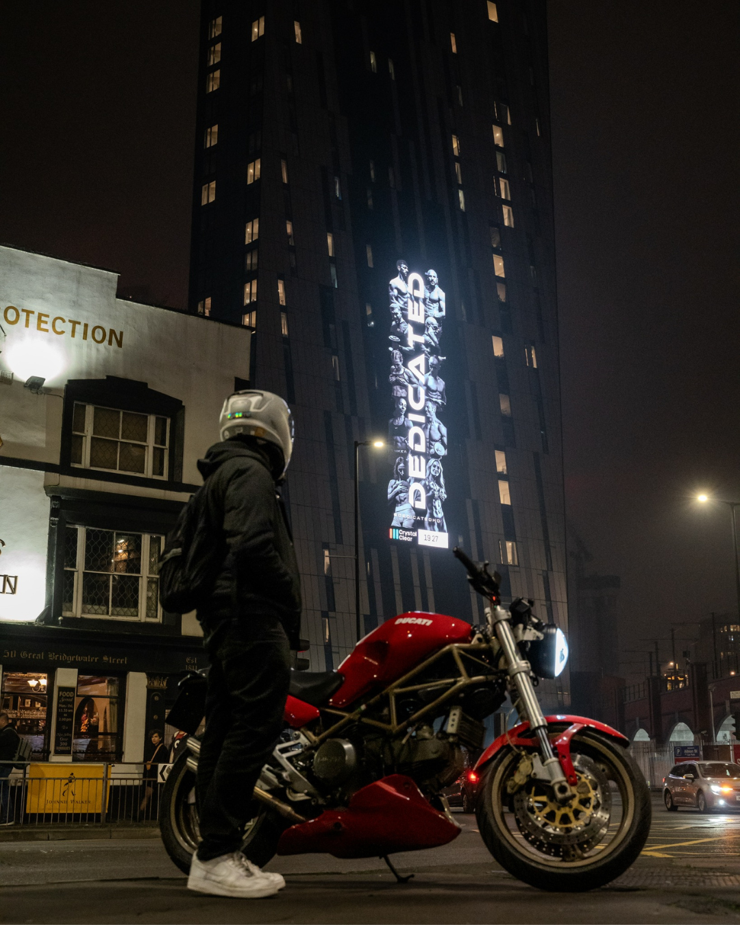 Person wearing a helmet and dark clothing standing next to a red Ducati motorcycle on a city street at night with a tall building in the background displaying a vertical illuminated advertisement of muscular athletes and the word DEDICATED.