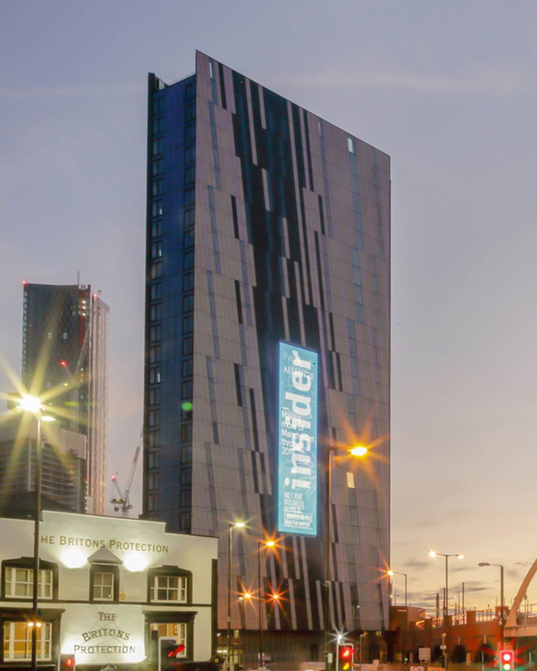 Tall modern building with patterned facade and bright vertical digital sign near a smaller white building named The Britons Protection at dusk.