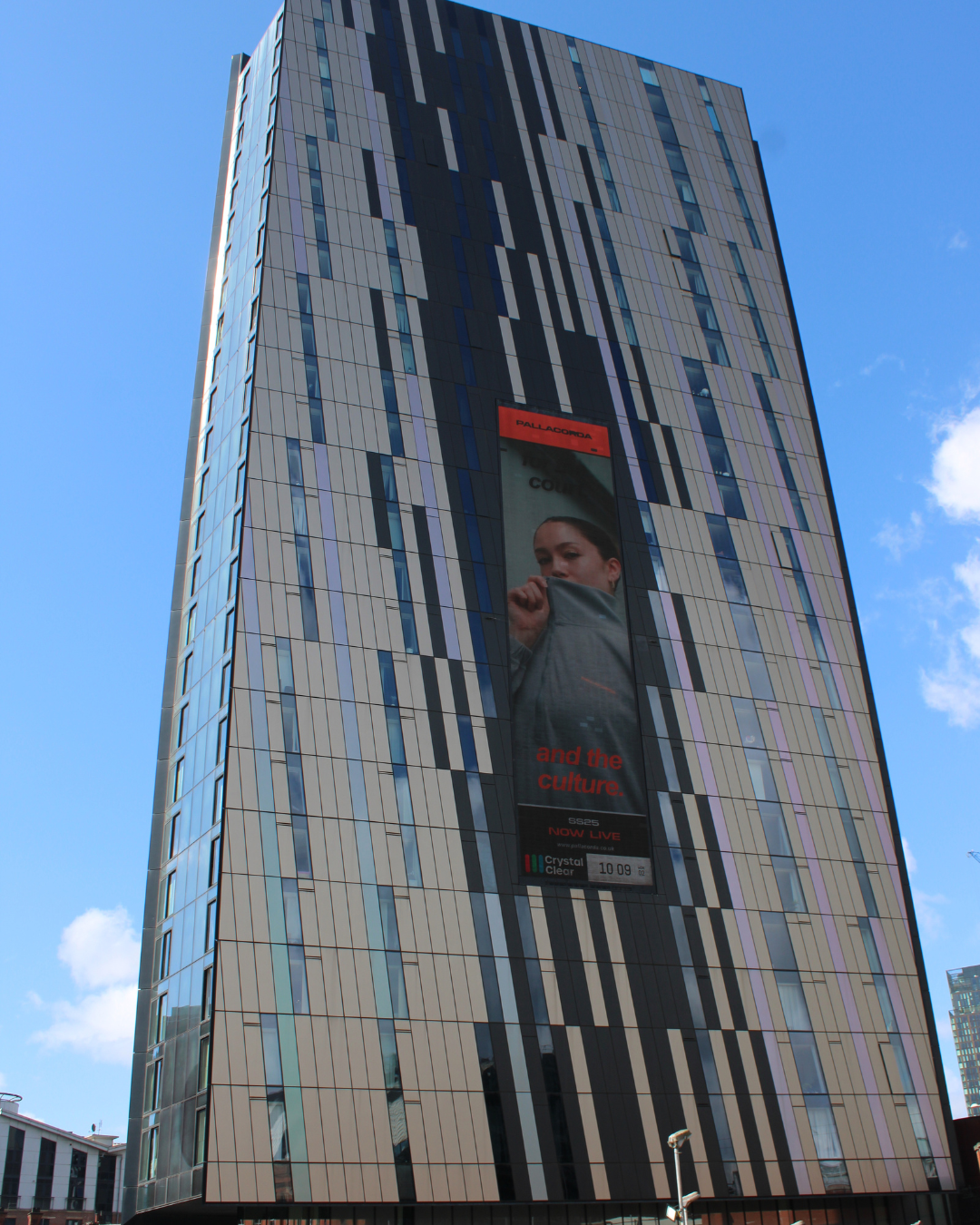 Tall modern building with vertical striped windows and a large vertical digital advertisement showing a woman partly covering her face.