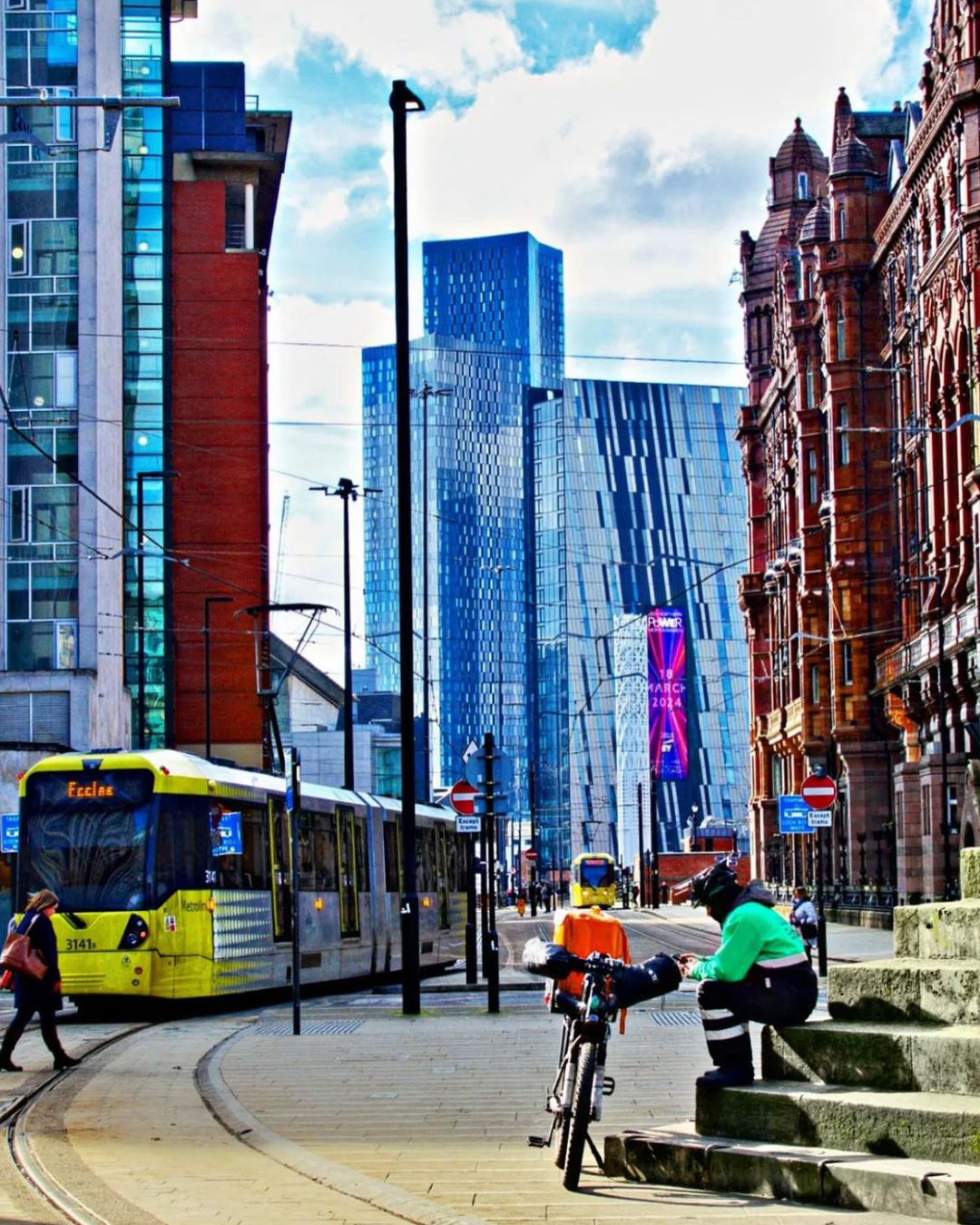 Urban street scene with a yellow tram on tracks, a cyclist resting on steps beside a loaded bike, and modern skyscrapers in the background.