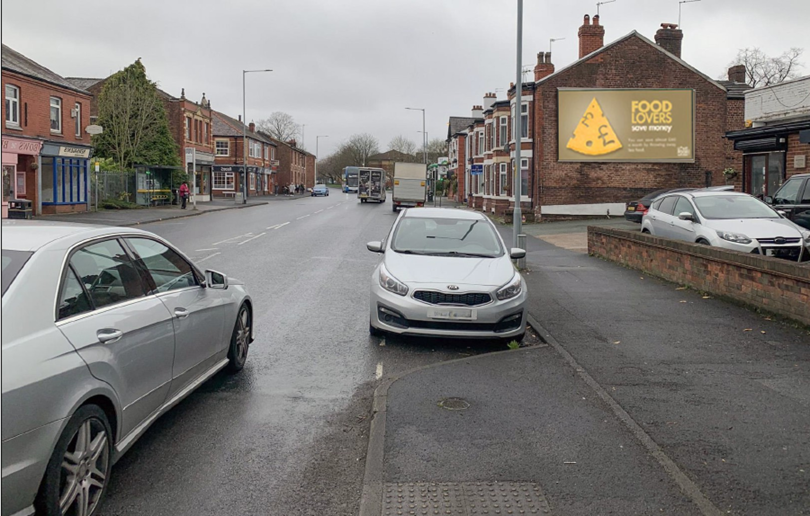 Overcast urban street with parked silver cars and a brick building featuring a billboard advertising food savings with a cheese wedge graphic.
