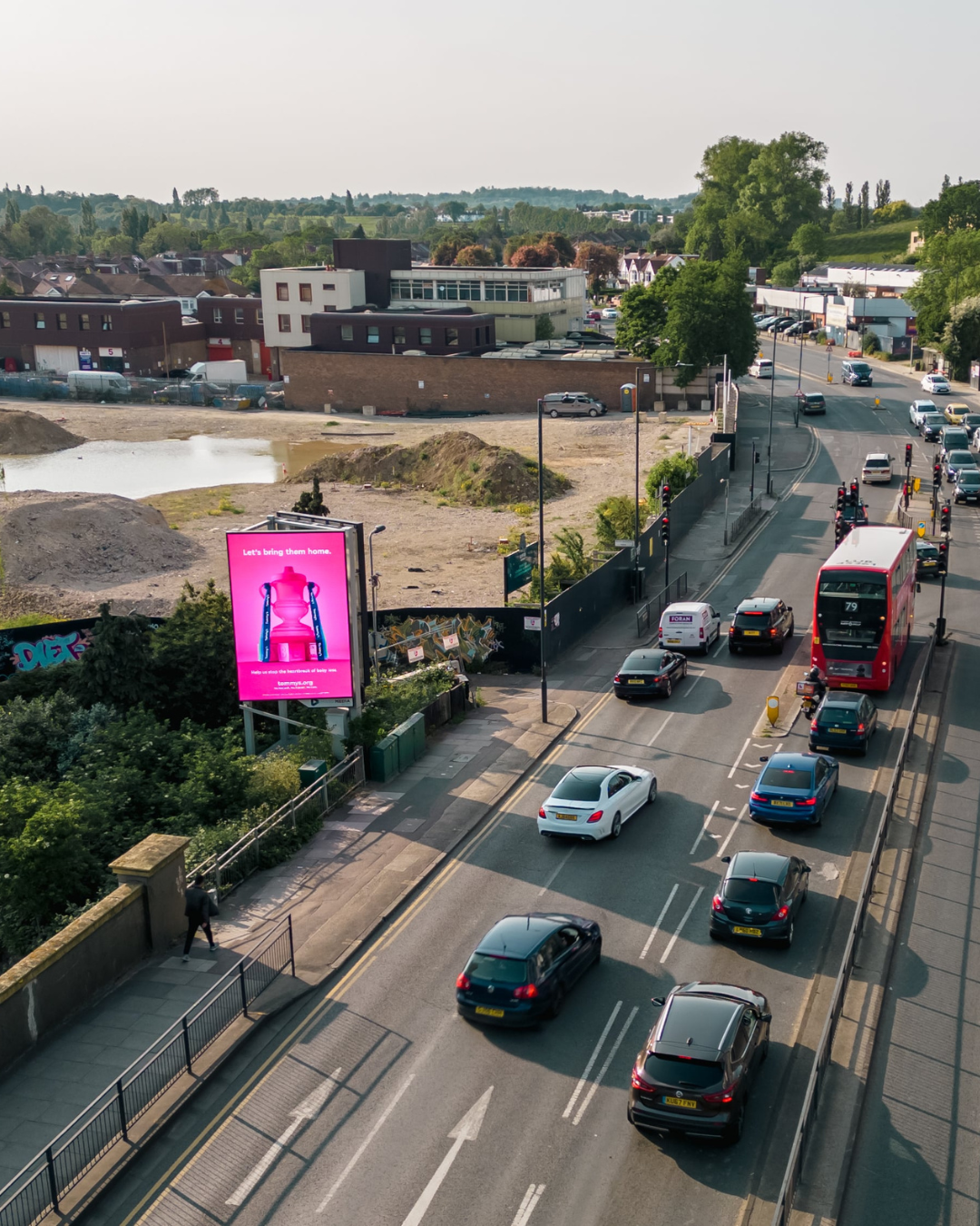 A busy street with cars and a red double-decker bus traveling past a billboard with a pink advertisement near an empty lot and buildings in the background.
