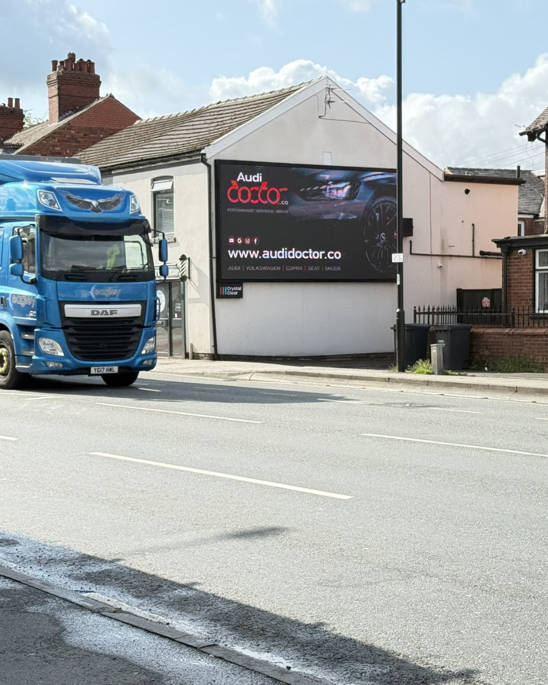 Blue DAF truck driving on a road past a white building with a large Audi Doctor advertisement on its wall.