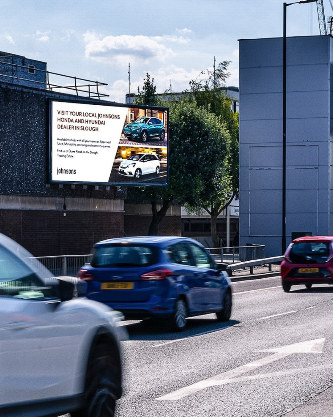 Cars driving on a street with a large billboard advertising Johnsons Honda and Hyundai dealer in Slough displayed on a building.