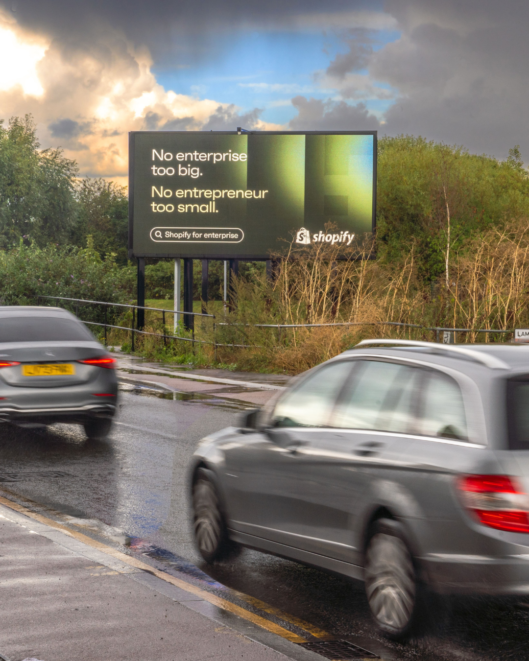 Outdoor billboard alongside a road with moving cars displaying a Shopify ad that reads, 'No enterprise too big. No entrepreneur too small.'