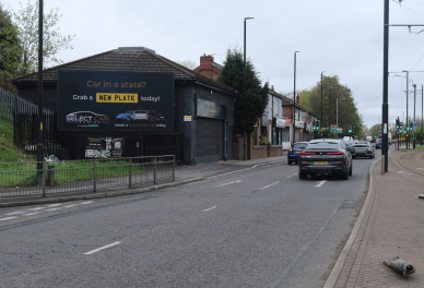 Street view with cars driving past a black billboard advertising new car plates, near a garage and shops on an overcast day.