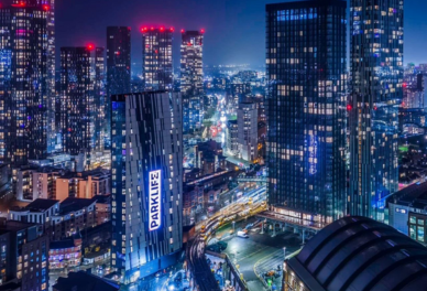 Nighttime cityscape with tall illuminated skyscrapers and a building displaying a vertical PARKLIFE sign.