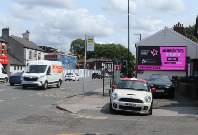 Street scene with parked white Mini Cooper and black car beside a building with a large pink digital billboard displaying 'Award Entries Now Open'.