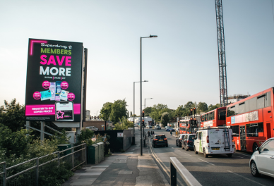 Urban street scene with a digital billboard advertising savings for members, surrounded by cars and red double-decker buses.