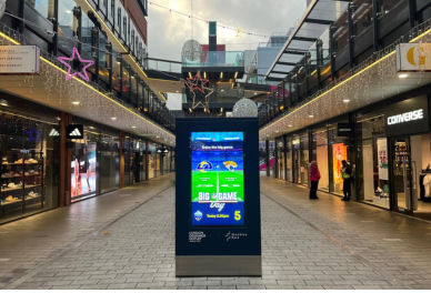 Indoor shopping street decorated with illuminated star and ball ornaments, with digital signage in center advertising 'Big Game' event.