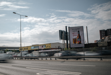 Urban street scene with moving cars, a billboard advertising beer, and industrial buildings under a partly cloudy sky.