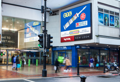 People walking near Reading Town Centre entrance with illuminated advertisements and storefronts on a rainy day.