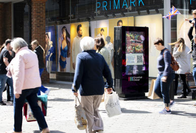 People walking past a Primark store with fashion posters in the window and a digital advertising kiosk on the sidewalk.