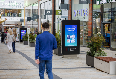 Man walking in a shopping mall corridor with digital voting information screens and multiple store signs in the background.