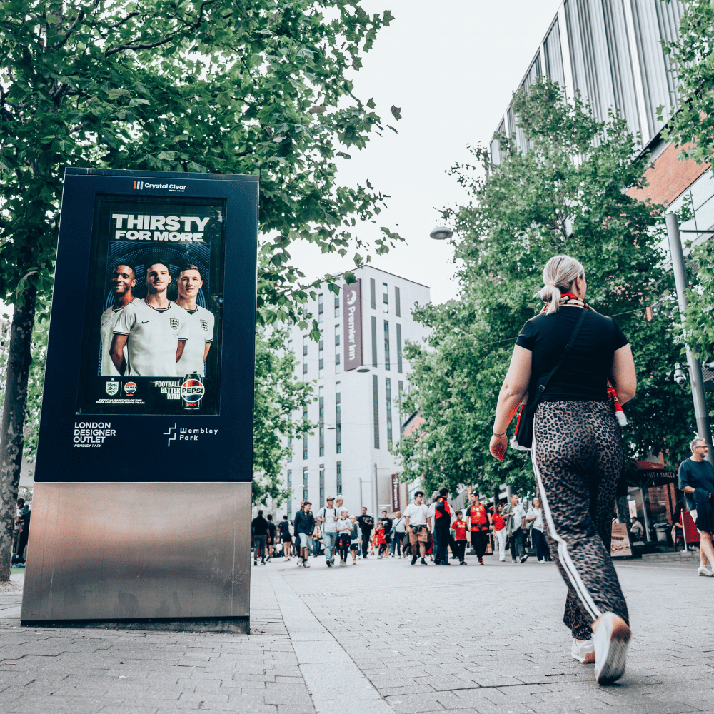 Outdoor scene at Wembley Park with a digital advertising board showing three men in white England soccer jerseys with text 'THIRSTY FOR MORE' and Pepsi branding, a woman walking away in leopard print pants and black shirt, and a crowd of people and trees along the paved street.