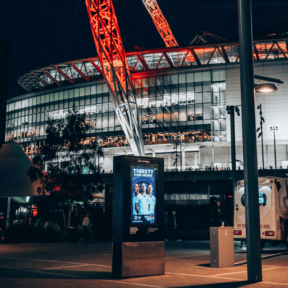 Night view outside Wembley Park with illuminated red steel supports of the stadium roof, glass windows showing interior lights, a digital advertisement board featuring three soccer players with the text 'Thirsty for More,' and an ice cream van parked nearby.